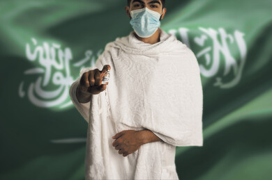 رجل مسلم عربي سعودي خليجي بلباس الاحرام ويرتدي الكمامة ،  اتباع تعليمات و شروط الحج لعام 2021 ، اخذ جرعة اللقاح الآمنة ، التطعيم الأمن لكوفيد 19  ، فريضة الحج ،  خلفية بعلم المملكة العربية السعودية