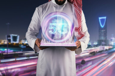 صورة مقربة لرجل عربي سعودي خليجي يقف أمام برج الرياض ، يحمل بيديه جهاز تقني ذكي عليه رسومات لمباني معمارية و مدنية ، يعلوها دائرة بلورية للكرة الأرضية بداخلها اختصار الجيل الخامس للشبكات 5G  بتقنية الهولوجرام ، استخدام خاصية الهولوجرام ثلاثية الأبعاد .