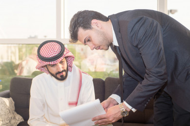 رجل اعمال سعودي عربي بلباس سعودي تقليدي  مع شريكه العربي بالبدلة ، يتناقشون على عقد عمل , اجتماع عمل ، شركة سعودية ، بيئة عمل