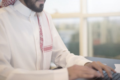 صورة مقربة لرجل اعمال سعودي خليجي على مكتب و يعمل على الكمبيوتر المحمول ، لباس السعودي التقليدي  ، شركة سعودية ، عمل الخليج ، وظيفة مكتبية ، بيئة عمل