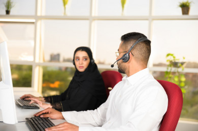 شابة موظفة عربية سعودية  و شاب موظف عربي سعودي يتحدثان في بيئة العمل , زملاء عمل ، العمل بالدعم الفني عبر الكمبيوتر 