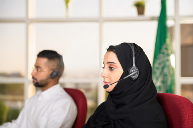 صورة مقربة لموظفة شابة عربية سعودية مع زميلها الموظف الشاب العربي السعودي في بيئة العمل , زملاء عمل ، العمل بالدعم الفني عبر الكمبيوتر
