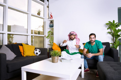 صديقان سعوديان يشاهدان مباراه للدوري السعودي ، كلاهما يشجعان فريق الأهلي وفريق السعودية ، ايماءات وجه مختلفة ، يجلسان على اريكة في غرفة المعيشة