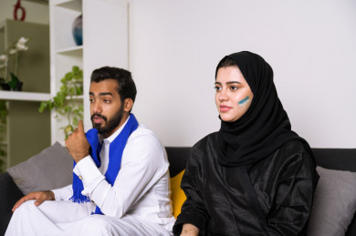 زوجان سعوديان يشاهدان مباراه للدوري السعودي ، الزوجة تشجع فريق الأهلي و السعودية ، و الزوج يشجع فريق الهلال
