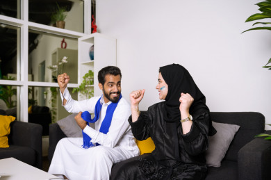 زوجان سعوديان يشاهدان مباراه للدوري السعودي ، الزوجة تشجع فريق الأهلي و السعودية ، و الزوج يشجع فريق الهلال