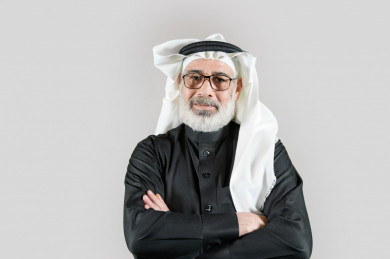 بورتريه رجل سعودي كبير بالسن يرتدي ثوب أسود مع غترة وعقال بأيدي متقاطعة بخلفية بيضاء