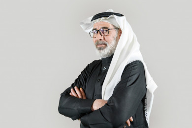 بورتريه رجل سعودي كبير بالسن يرتدي ثوب أسود مع غترة وعقال بأيدي متقاطعة بخلفية بيضاء