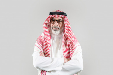 بورتريه رجل سعودي كبير بالسن يرتدي ثوب أبيض مع شماغ وعقال بأيدي متقاطعة بخلفية بيضاء