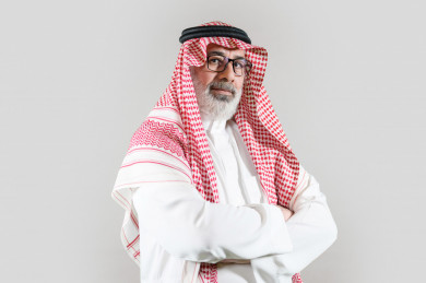 بورتريه رجل سعودي كبير بالسن يرتدي ثوب أبيض مع شماغ وعقال بأيدي متقاطعة بخلفية بيضاء