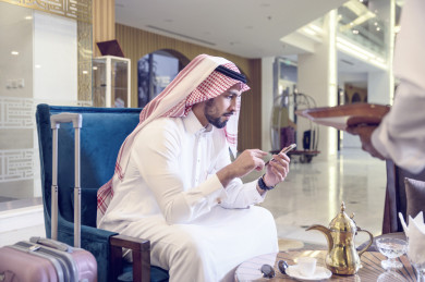 نزيل سعودي يجلس في منطقة الانتظار في بهو فندق ويستخدم الهاتف الجوال بجانبه شنطة سفر 