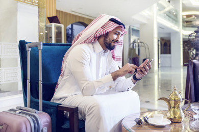 نزيل سعودي يجلس في منطقة الانتظار في بهو فندق ويستخدم الهاتف الجوال بجانبه شنطة سفر 