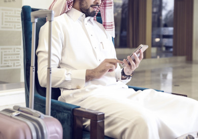 صورة مقربة لنزيل سعودي يجلس في منطقة الانتظار في بهو فندق ويستخدم الهاتف الجوال بجانبه شنطة سفر 