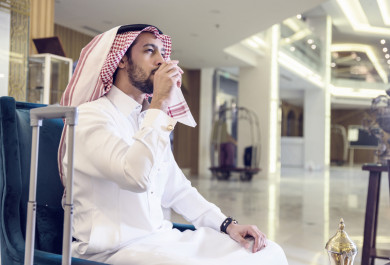 نزيل سعودي يجلس في منطقة الانتظار في بهو فندق ويشرب القهوة بجانبه شنطة سفر 
