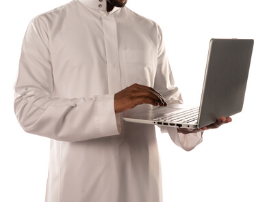 بورتريه لشاب أسمر عربي خليجي سعودي يحمل بيده جهاز حاسوب محمول، انجاز مهام من خلال الحاسوب المحمول، العمل عن بعد، أجهزة تقنية حديثة، استخدام تقنيات حديثة، بشرة سمراء، خلفية بيضاء