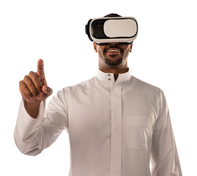 بورتريه لشاب أسمر عربي خليجي سعودي يرتدي نظارة الواقع الإفتراضي ثلاثية الأبعاد، يشير بأصبع السبابة لشئ ما يشاهده بالواقع الإفتراضي، إستخدام حاسة اللمس، محاكاة العالم الافتراضي، خلفية بيضاء
