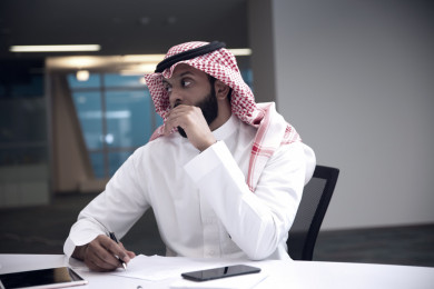 صورة مقربة رجل اعمال سعودي خليجي على المكتب باجتماع  مع فريق العمل ، يسجل ملاحظات بالقلم ، شركة سعودية ، باللباس السعودي التقليدي ، بيئة عمل 