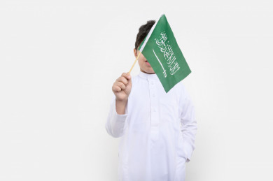 بورتريه فتى عربي سعودي يمسك علم السعودية بيده ، يرفرف بيده علم السعودية ، اليوم الوطني السعودي ، بخلفية بيضاء