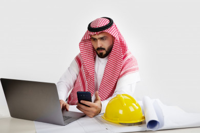 بورتريه مهندس معماري سعودي خليجي محترف في العمل ، يتابع العمل على الكمبيوتر المحمول ، يخطط لمشروع جديد , مهندس انشاءات خليجي بالثوب السعودي ، مخطط البناء ، خوذة العمل ، خلفية بيضاء
