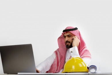 بورتريه مهندس معماري سعودي خليجي محترف في العمل ، يتابع العمل على الكمبيوتر المحمول ، يخطط لمشروع جديد , مهندس انشاءات خليجي بالثوب السعودي ، مخطط البناء ، خوذة العمل ، خلفية بيضاء