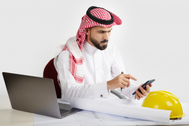بورتريه مهندس معماري سعودي خليجي في مكتب العمل ، استخدام الهاتف المحمول ، يخطط لمشروع جديد , مهندس انشاءات خليجي بالثوب السعودي ، مخطط البناء ، خوذة العمل ، خلفية بيضاء