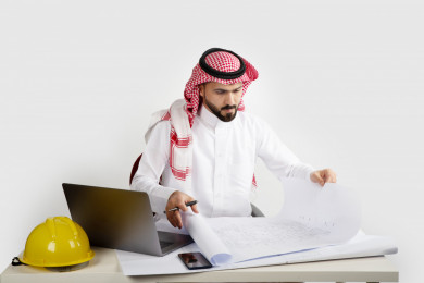 بورتريه مهندس معماري سعودي خليجي محترف  في مكتب العمل ، يتابع العمل على مخططات البناء ، يخطط لمشروع جديد , مهندس انشاءات خليجي بالثوب السعودي ، مخطط البناء ، خوذة العمل ، خلفية بيضاء 