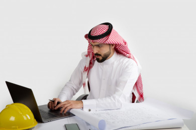 بورتريه مهندس معماري سعودي خليجي في العمل  ، يخطط لمشروع جديد على الكمبيوتر المحمول , مهندس انشاءات خليجي بالثوب السعودي ، مخطط البناء ، خوذة العمل ، خلفية بيضاء 