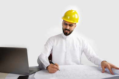 بورتريه مهندس معماري سعودي  مبتسم على مكتبه مخطط البناء ، يسجل بيانات على  ورقة تخطيط البناء ، مخطط البناء ، خوذة  عمل صفراء ، مهندس انشاءات خليجي ، خلفية بيضاء