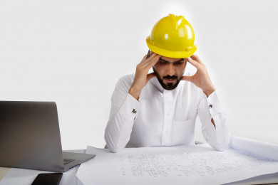 بورتريه مهندس معماري سعودي على مكتبه مخطط البناء ، يواجه التفكير الصعب بالمشروع الجديد  ، مخطط البناء ، خوذة  عمل صفراء ، مهندس انشاءات خليجي ،خلفية بيضاء