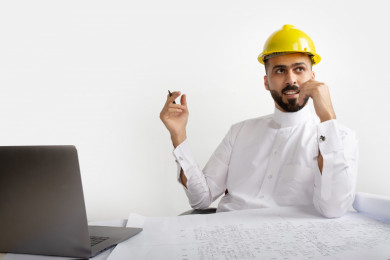بورتريه مهندس معماري سعودي على مكتبه مخطط البناء ، يفكر بالمشروع الجديد ، مخطط البناء ، خوذة  عمل صفراء ، مهندس انشاءات خليجي ، خلفية بيضاء