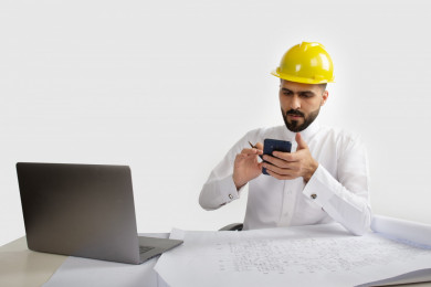 بورتريه مهندس معماري سعودي على مكتبه مخطط البناء ، بيده الهاتف المحمول ، مخطط البناء ، خوذة  عمل صفراء ،مهندس انشاءات خليجي ، خلفية بيضاء