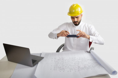 بورتريه مهندس معماري سعودي على مكتبه مخطط البناء ، يلتقط صور لمخطط البناء باستخدام الهاتف المحمول ، مخطط البناء ، خوذة  عمل صفراء ، مهندس انشاءات خليجي ، خلفية بيضاء