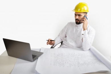 بورتريه مهندس معماري سعودي في العمل ، يجلس على مكتبه ويتحدث بالتلفون المحمول  ، يخطط لمشروع جديد على الكمبيوتر المحمول ، مخطط البناء ، خوذة  عمل صفراء ، مهندس انشاءات خليجي ، خلفية بيضاء