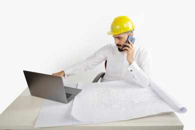 بورتريه مهندس معماري سعودي في العمل ، يجلس على مكتبه ويتحدث بالتلفون المحمول  ، يخطط لمشروع جديد على الكمبيوتر المحمول ، مخطط البناء ، خوذة  عمل صفراء ، مهندس انشاءات خليجي ،خلفية بيضاء
