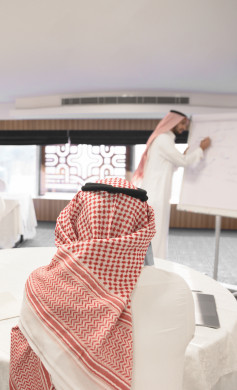 مدرب عربي سعودي يقوم بالشرح لمتدرب سعودي خليجي على سبورة بيضاء ، رجل اعمال سعودي يتحدث عن  استراتيجيات الشركة على لوحة بيضاء الى الزملاء في العمل ، دورات تدريبية ، متدربين ، اللباس السعودي التقليدي ، غرفة اجتماعات ، اجتماع عمل