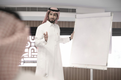 مدرب عربي سعودي يقوم بالشرح لمتدربين سعوديين خليجيين على سبورة بيضاء ، رجل اعمال سعودي يتحدث عن  استراتيجيات الشركة على لوحة بيضاء الى الزملاء في العمل ، دورات تدريبية ، متدربين ، اللباس السعودي التقليدي ، غرفة اجتماعات ، اجتماع عمل