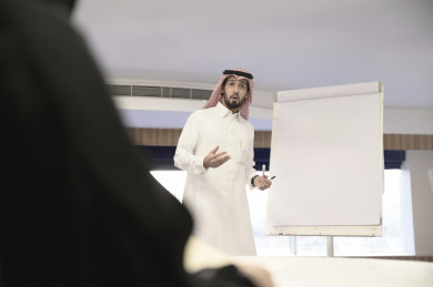 مدرب عربي سعودي يقوم بالشرح لمتدربين سعوديين خليجيين على سبورة بيضاء ، رجل اعمال سعودي يتحدث عن  استراتيجيات الشركة على لوحة بيضاء الى الزملاء في العمل ، دورات تدريبية ، متدربين ، اللباس السعودي التقليدي ، غرفة اجتماعات ، اجتماع عمل
