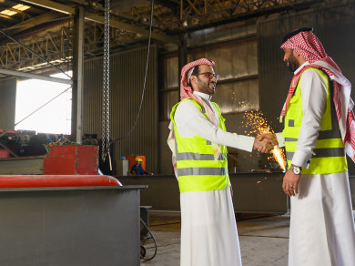 مهندسان صناعيان سعوديين شركاء في مصنع الحديد ، يتصافحان لبدء مشروع جديد ، باللباس السعودي التقليدي ،شرارة لحام الحديد مبعثرة بالخلفية ، مفهوم الهندسة و الصناعة