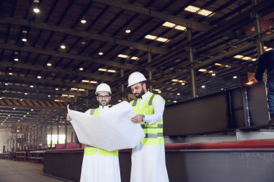 مهندسان صناعيان سعوديان  يشرفان على المصنع ، يحملان  ورقة مخطط المصنع  ، ارتداء خوذة  و سترة الحماية الخاصة بالعمل