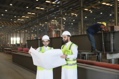 مهندسان صناعيان سعوديان  يشرفان على المصنع ، يحملان  ورقة مخطط المصنع  ، ارتداء خوذة  و سترة الحماية الخاصة بالعمل