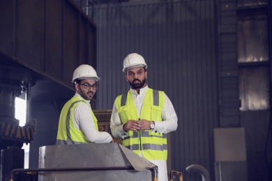 مهندسان صناعيان سعوديين شركاء في مصنع الحديد ، يمسكان بيد الأخر ، احدهم يؤشر بيده نحو اماكن الانتاج المستقبليه