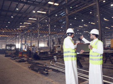 مهندسان سعوديان داخل مصنع الحديد ، يفحصان جودة الحديد من خلال الكمبيوترالمحمول ، ارتداء خوذة و سترة الحماية الخاصة بالعمل ، مفهوم الهندسة و الصناعة
