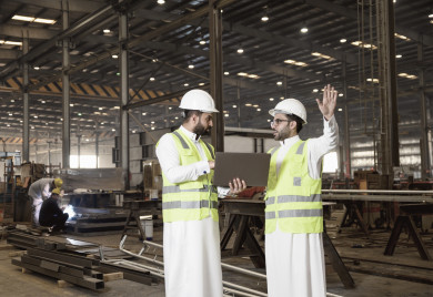 مهندسان سعوديان داخل مصنع الحديد ، يفحصان جودة الحديد من خلال الكمبيوترالمحمول ، ارتداء خوذة و سترة الحماية الخاصة بالعمل ، مفهوم الهندسة و الصناعة