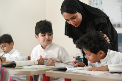معلمة خليجية تشرف على طالب عن قرب وتمسح على رأسه, مدرسة سعودية تنظر إلى كتاب طالب وتبتسم, أستاذة عربية تقرأ حل طالب وتربت على رأسه