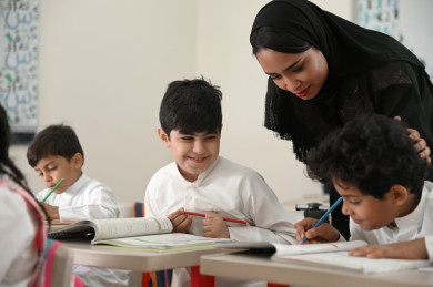 معلمة خليجية تشرف على طالب عن قرب وتمسح على رأسه, مدرسة سعودية تنظر إلى كتاب طالب وتبتسم, أستاذة عربية تقرأ حل طالب وتربت على رأسه