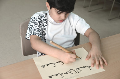 طفل سعودي يكتب جملة همة حتى القمة بالخط العربي، طالب خليجي يتدرب على الكتابة في درس الخط العربي 