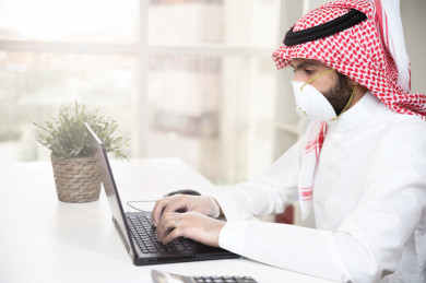 رجل اعمال سعودي خليجي يرتدي الكمامة والقفازات لضبط انتشار العدوى ، استخدام الكمبيوتر المحمول لانجاز العمل ، العمل عن بعد من المنزل ، اجتماعات اونلاين