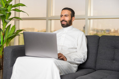 رجل اعمال سعودي خليجي يعمل عن بعد في المنزل  لضبط انتشار العدوى ، استخدام الكمبيوتر المحمول لانجاز العمل ، يشعر بالسعادة لانجاز العمل بنجاح عن بعد ، اجتماعات اونلاين