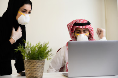 رجل اعمال سعودي خليجي يرتدي الكمامة والقفازات لضبط انتشار العدوى ،بجانبه السكرتيرة تتبع اجراءات السلامة و الوقاية ايضا ، استخدام الكمبيوتر المحمول لانجاز العمل 