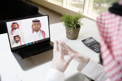 رجل اعمال سعودي خليجي يرتدي الكمامة والقفازات لضبط انتشار العدوى ، استخدام الكمبيوتر المحمول لانجاز العمل ، العمل عن بعد من المنزل ، اجتماعات اونلاين