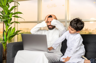 رجل اعمال سعودي خليجي يعمل عن بعد في المنزل  لضبط انتشار العدوى ، استخدام الكمبيوتر المحمول لانجاز العمل ، يشعر بالانزعاج بسبب تشويش الابن له اثناء العمل ، اجتماعات اونلاين